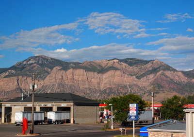 [Blue skies with patchy clouds are above the green tipped (from the trees) Wasatch mountains which extend well above the buildings in the foreground.]